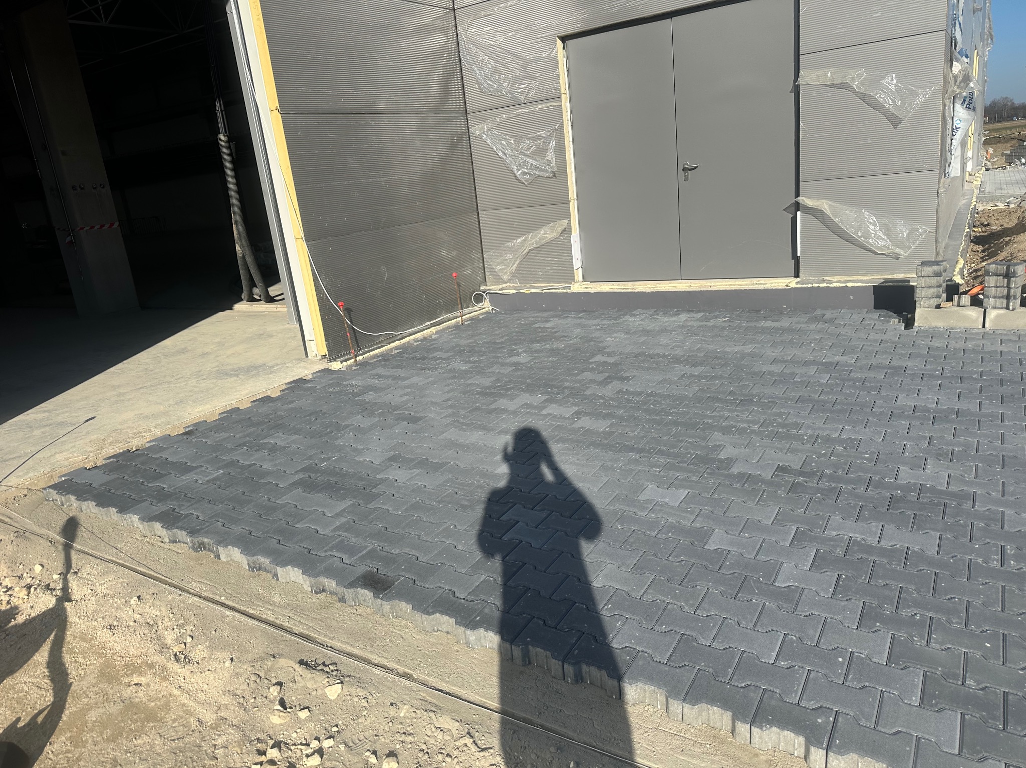 Fragment nowej, szarej nawierzchni z kostki brukowej ułożonej przed budynkiem przemysłowym z szarymi drzwiami, w tle widoczne materiały budowlane i cień osoby robiącej zdjęcie.