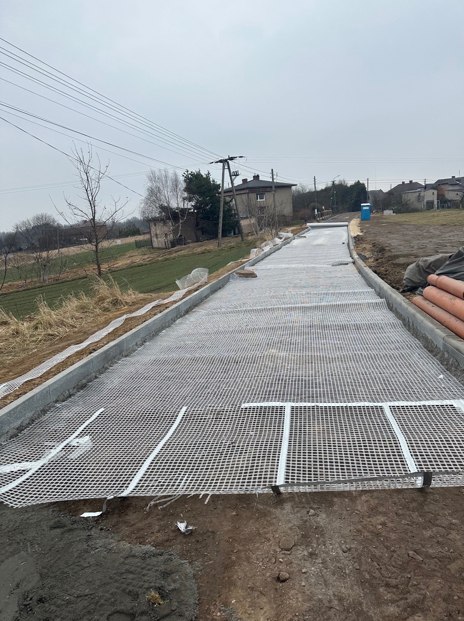 Przygotowanie podłoża pod nawierzchnię drogi z zastosowaniem siatki wzmacniającej, widoczne krawężniki i rury drenarskie, w tle zabudowa wiejska i pochmurne niebo.