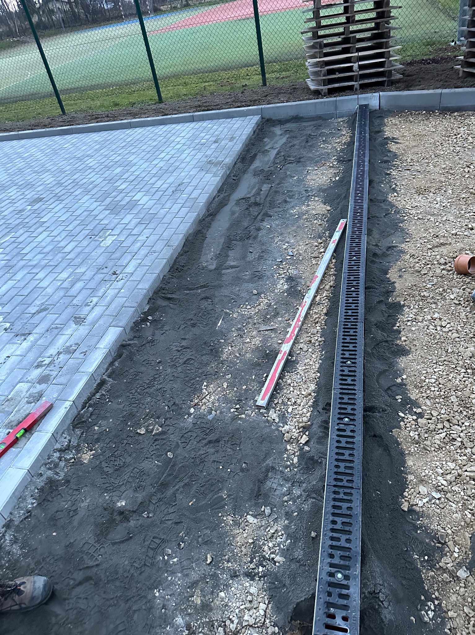Fragment placu z ułożoną szarą kostką brukową oraz przygotowanym podłożem z zamontowanym liniowym odpływem wody, widoczne poziomice i palety w tle.