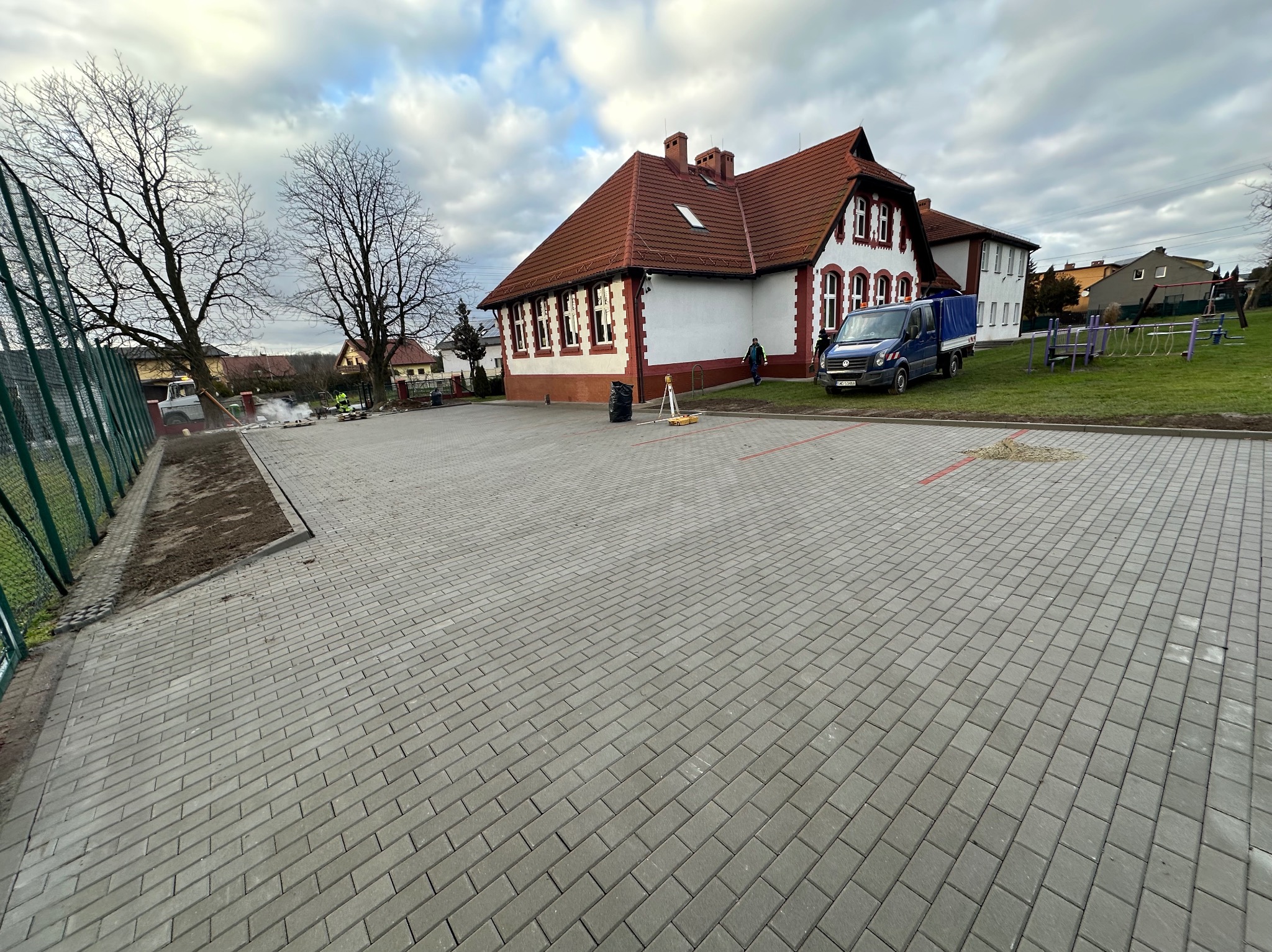 Nowa, szara nawierzchnia z kostki brukowej na tle budynku szkoły z czerwoną dachówką i zaparkowanym niebieskim samochodem dostawczym.
