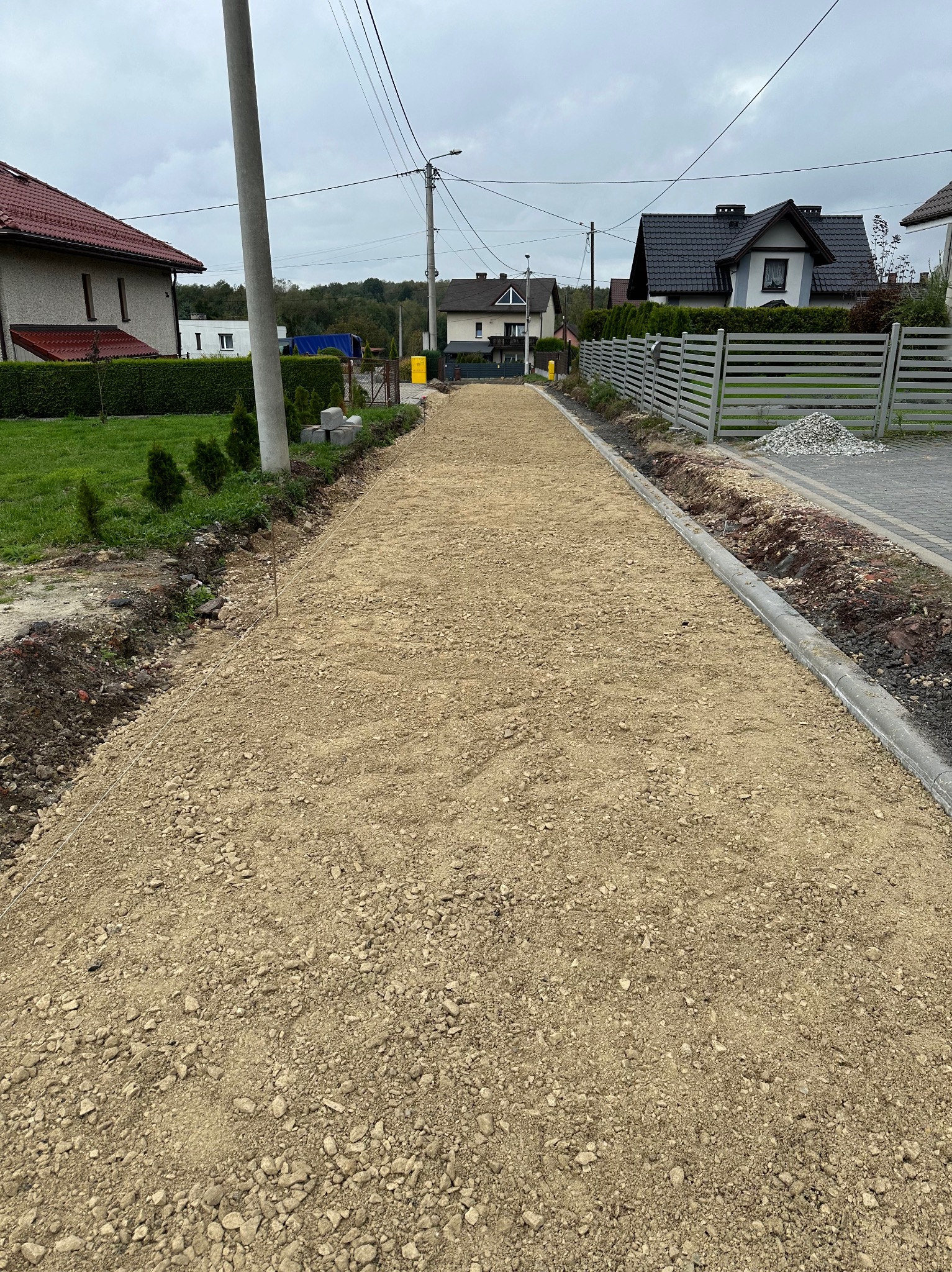 Przygotowanie podłoża z podsypką piaskowo-kamienną do ułożenia nawierzchni na posesji, widoczne obrzeża i wytyczone sznurki. W tle domy jednorodzinne i słupy energetyczne.