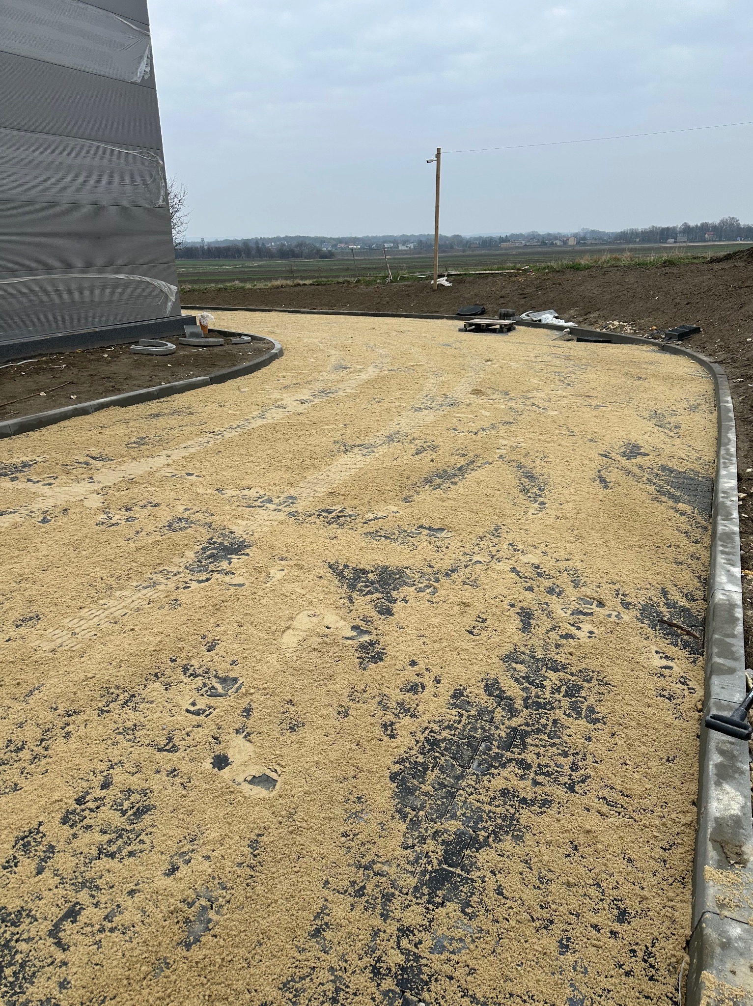 Przygotowanie podłoża pod nawierzchnię z kostki brukowej: wyrównany piasek na ułożonej kostce, obrzeża betonowe, widoczne ślady opon.