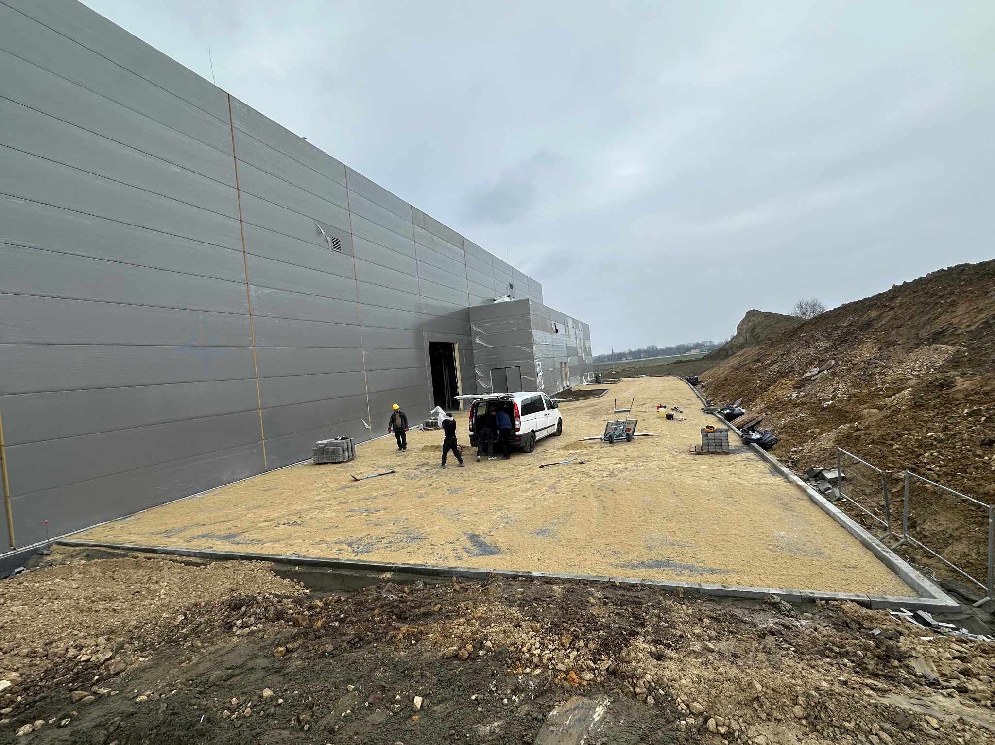 Przygotowanie podłoża piaskiem pod kostkę brukową przed budynkiem magazynowym, widoczni pracownicy i samochód dostawczy, obrzeża chodnika wykończone