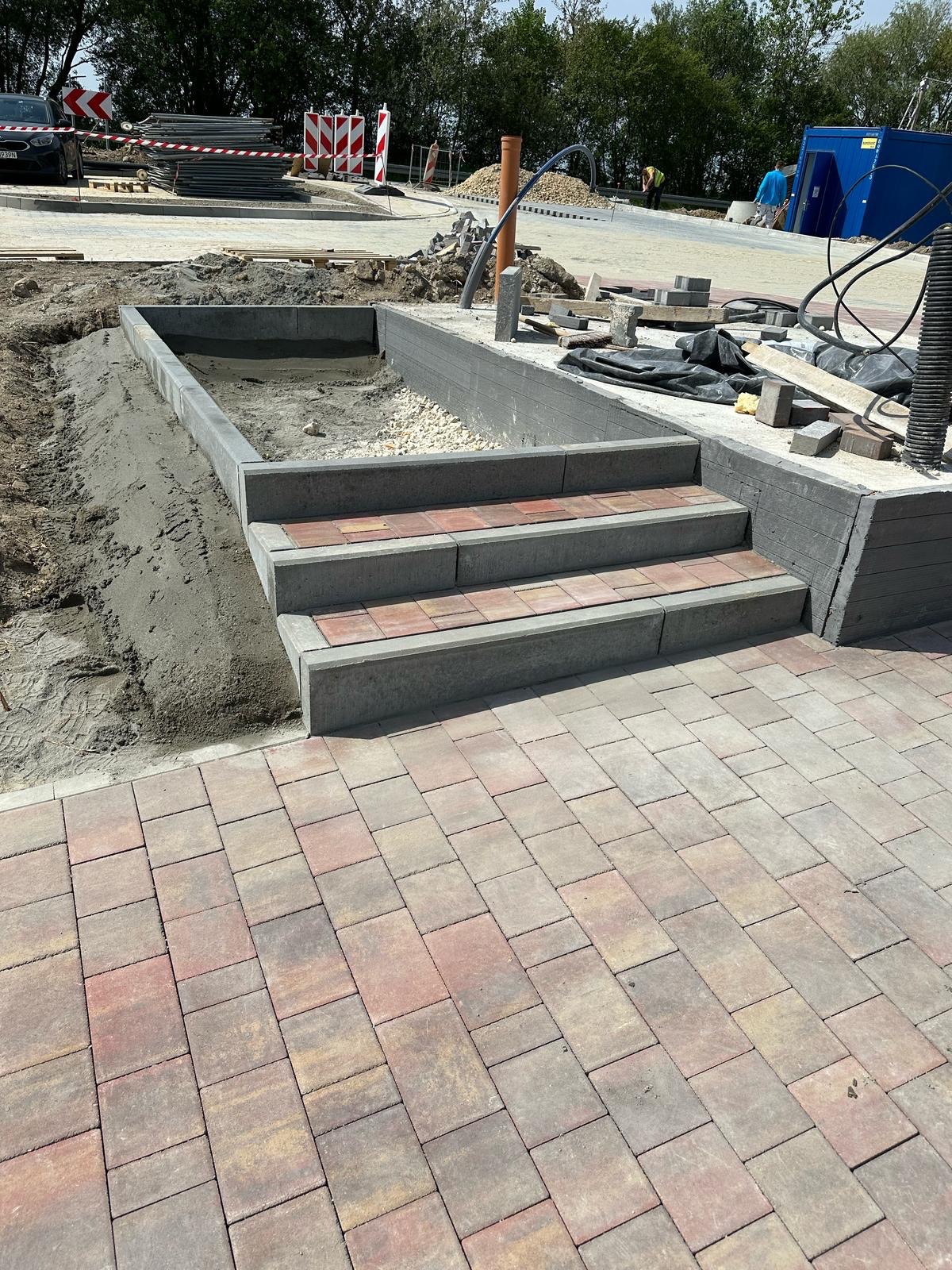 Wykończone schody z kostki brukowej prowadzące do podwyższonego rabatu, otoczone świeżo ułożoną nawierzchnią z kostki w odcieniach czerwieni i szarości. W tle plac budowy.