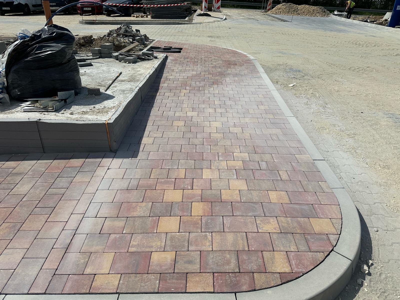 Ułożona kostka brukowa w odcieniach czerwieni i beżu, tworząca ścieżkę z zaokrąglonym brzegiem. W tle widoczne materiały budowlane i teren przygotowany pod dalsze prace.