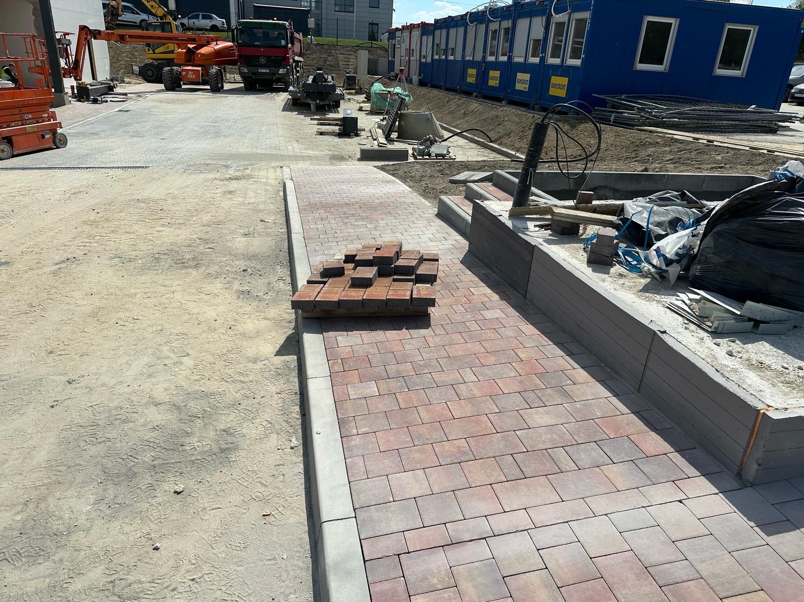Częściowo ułożona kostka brukowa w Połomi. Widoczne maszyny budowlane i baraki. Kostka w odcieniach czerwieni i szarości.