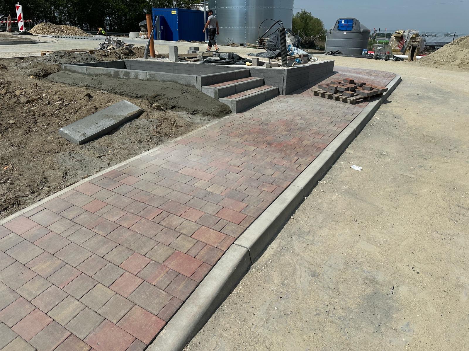 Układanie kostki brukowej: ścieżka z kostki w odcieniach czerwieni i brązu, obrzeża betonowe, schody z kostki, elementy przygotowane do ułożenia, teren budowy.