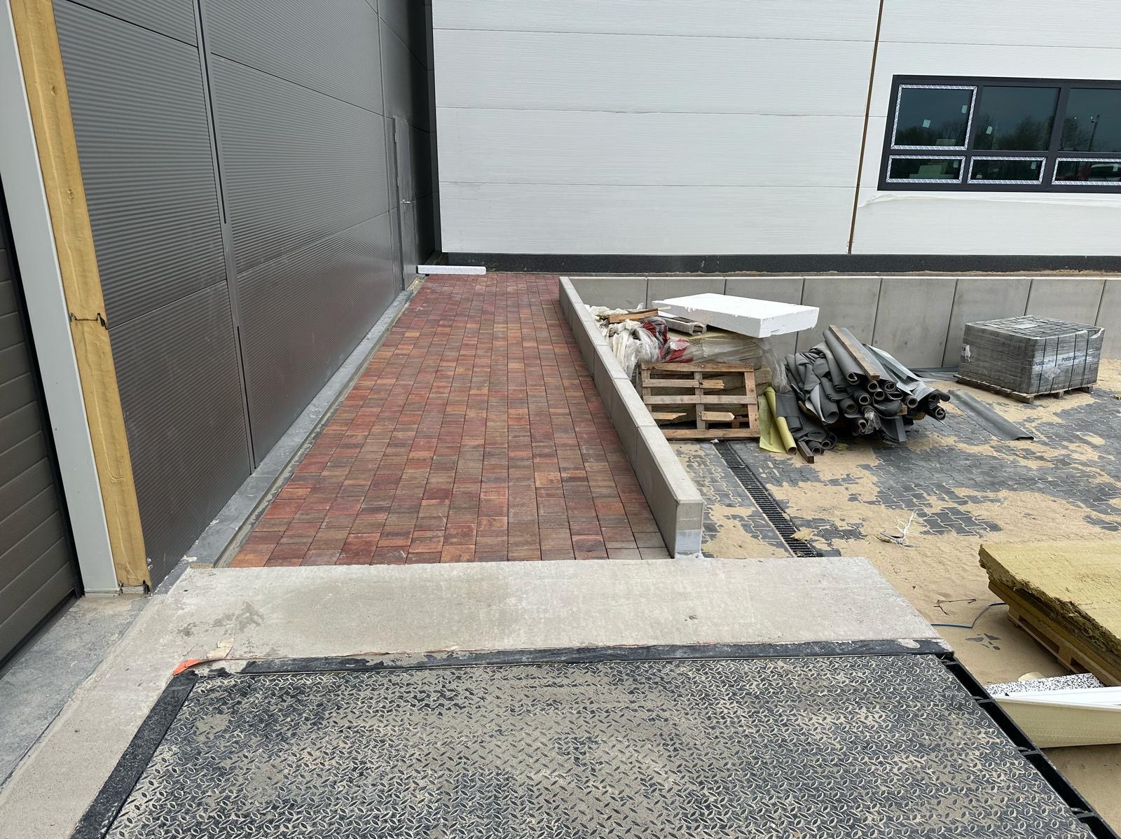 Ceglana ścieżka z kostki brukowej prowadzi do budynku przemysłowego, obok paleta z materiałami budowlanymi i rurami. Widoczny drenaż liniowy.