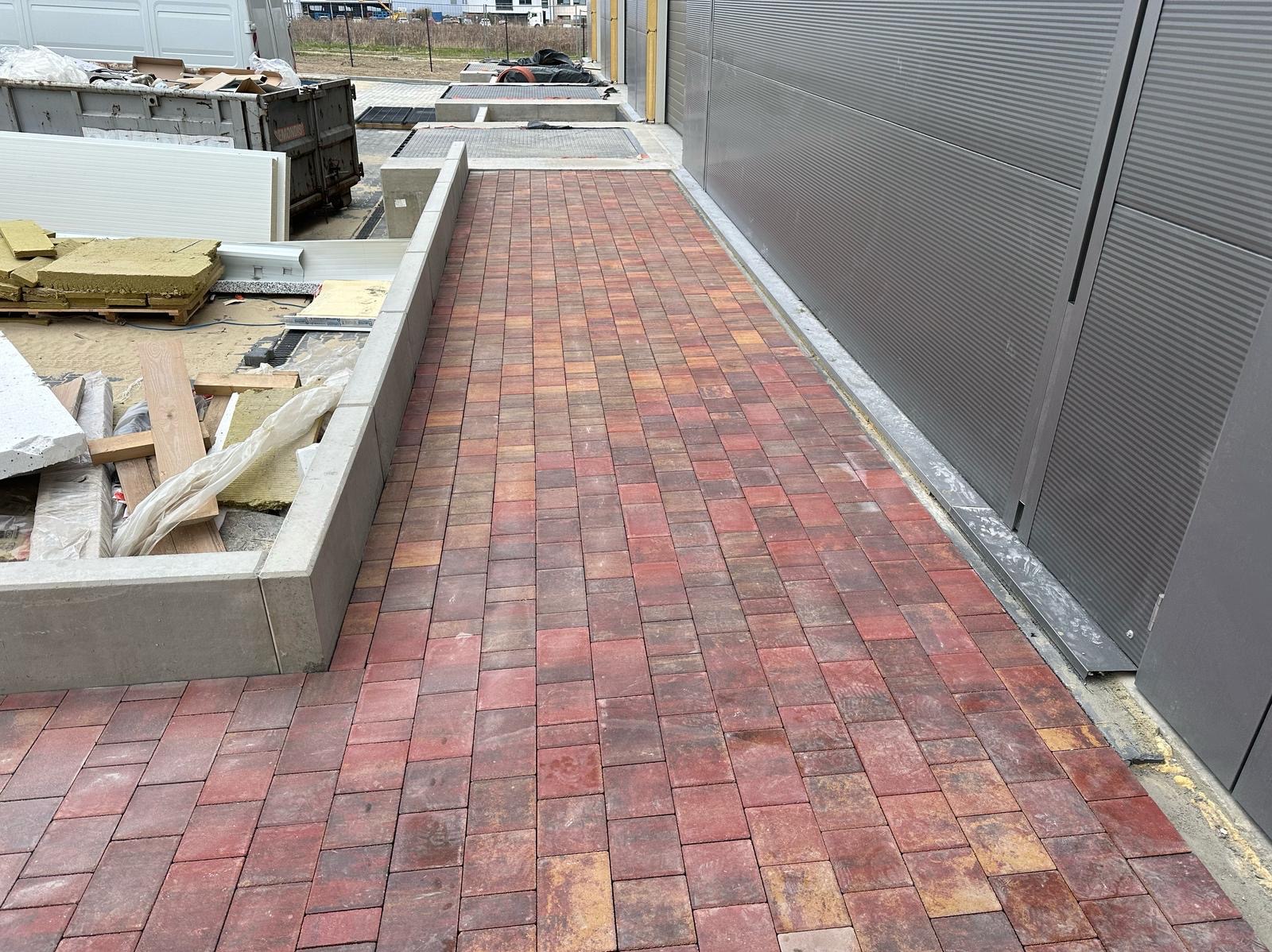 Ścieżka z czerwono-brązowej kostki brukowej, ułożona wzdłuż szarej, metalowej ściany budynku. Widoczne elementy placu budowy i betonowe obrzeże.