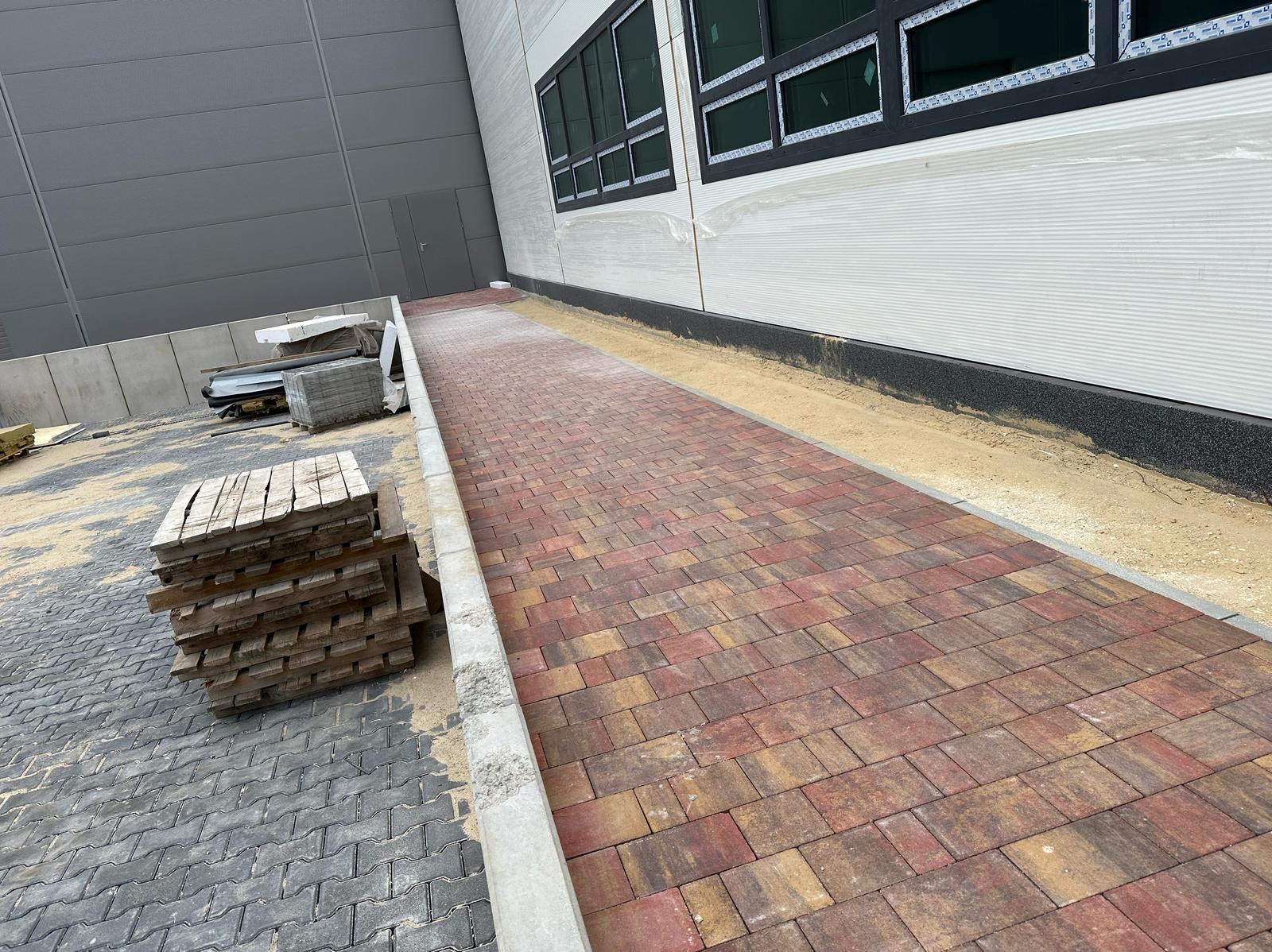 Kaskada kolorowej kostki brukowej prowadząca wzdłuż budynku z szarymi panelami. Paleta z kostką i fragment szarej kostki po lewej stronie.