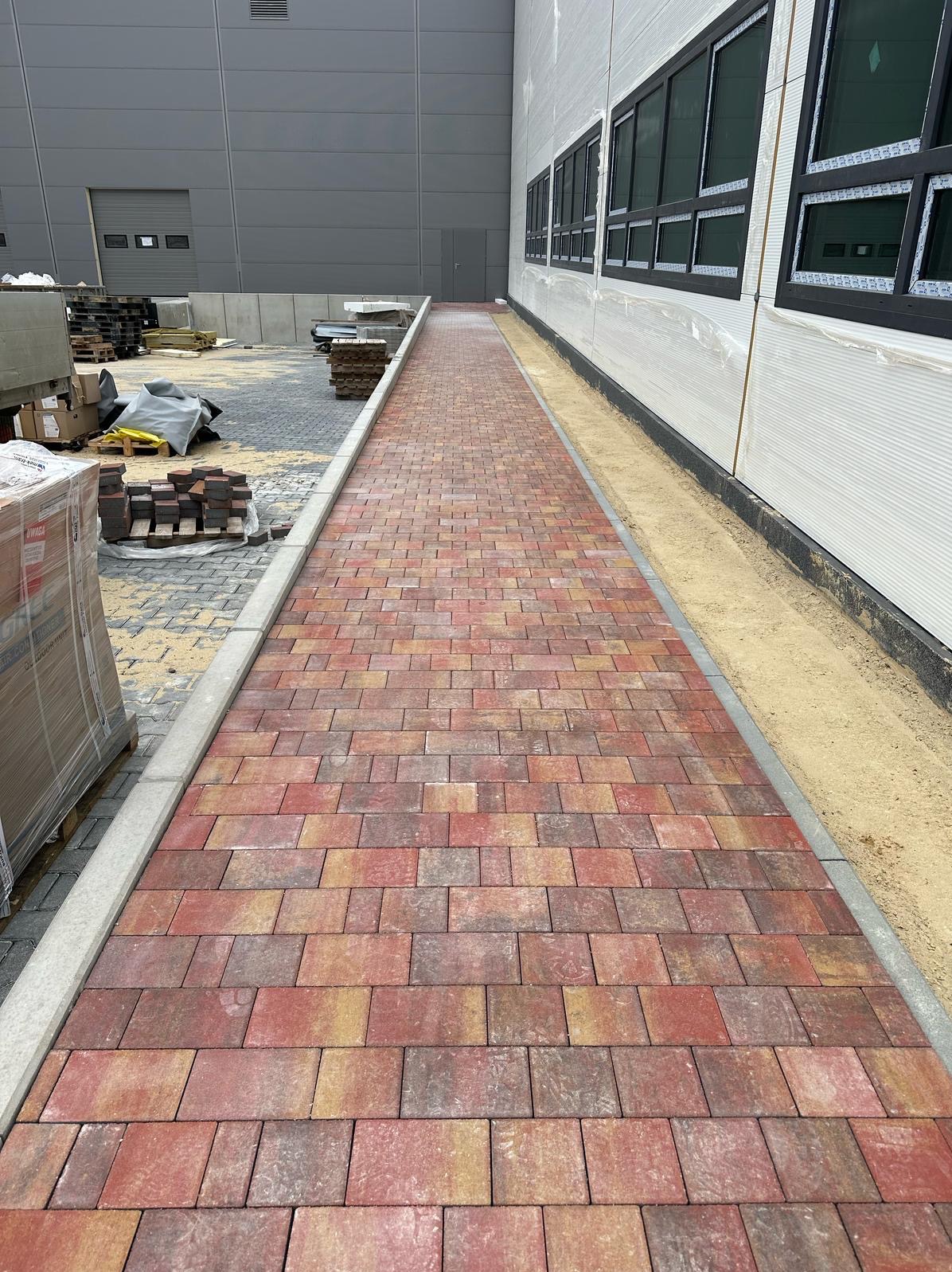 Czerwono-żółta kostka brukowa ułożona wzdłuż budynku przemysłowego, ograniczona szarym obrzeżem. Widoczne palety z kostką w tle.