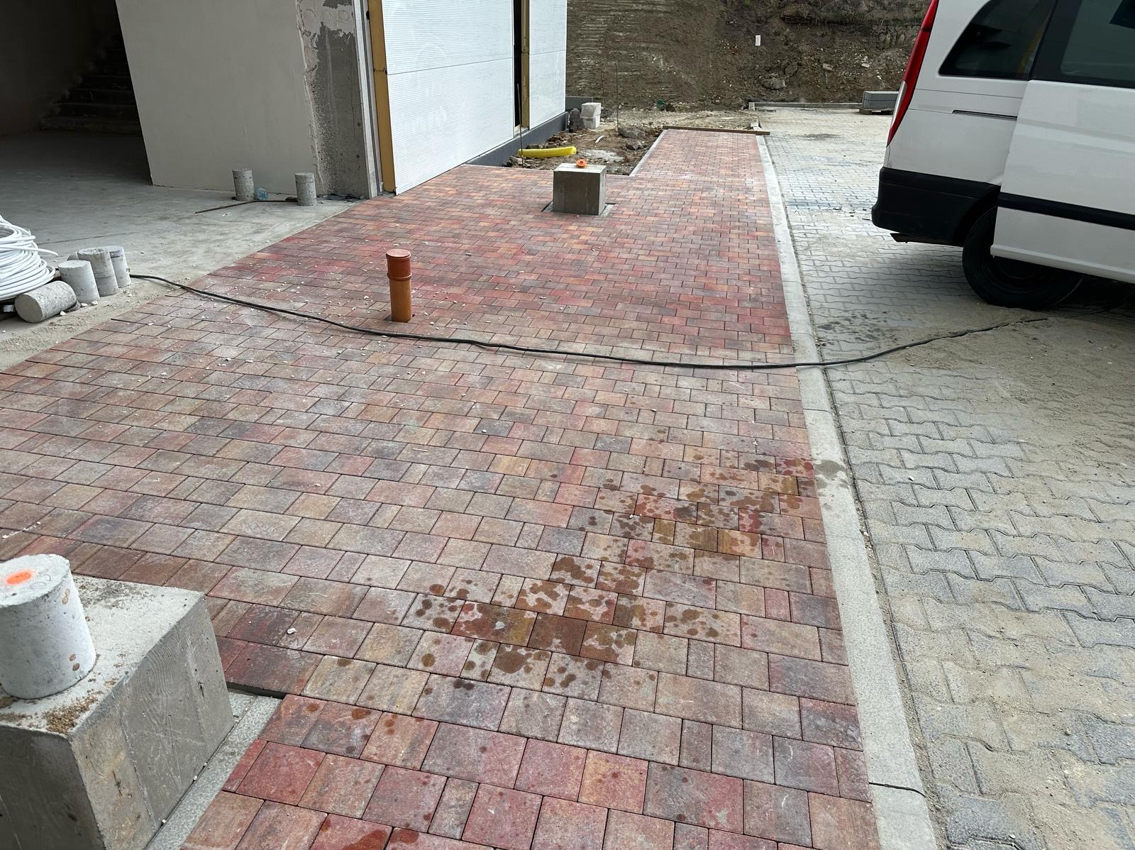 Częściowo ułożona, czerwono-brązowa kostka brukowa przed budynkiem z betonowymi elementami i zaparkowanym białym vanem. Widoczne ślady po wodzie.