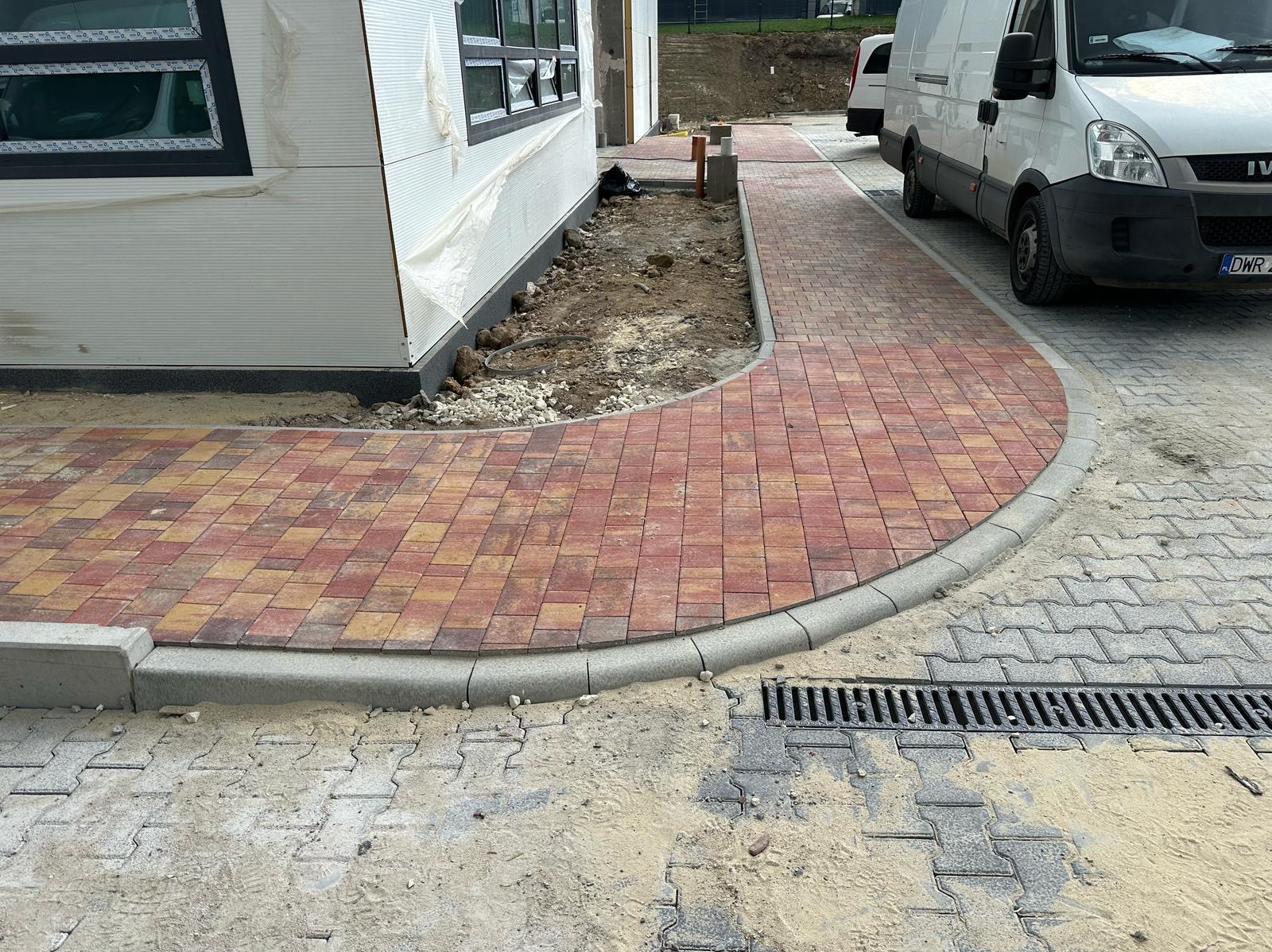 Czerwono-żółta kostka brukowa ułożona w łuk, obrzeża z szarego betonu, studzienka drenażowa, obok budynek w trakcie budowy i biały bus.