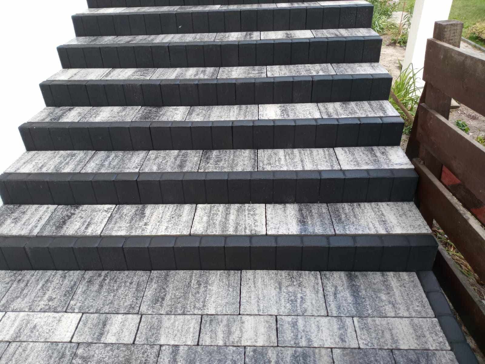 Schody z kostki brukowej w odcieniach szarości i czerni, z wyraźnie zaznaczonymi krawędziami stopni, prowadzące w górę w otoczeniu zieleni i drewnianego płotu.