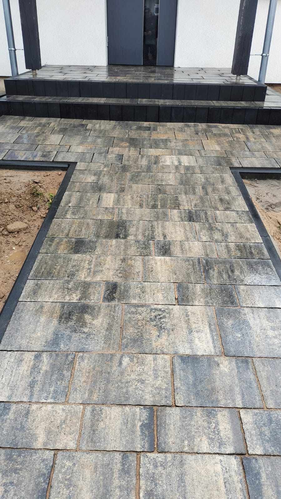 Ułożona, grafitowo-beżowa kostka brukowa prowadząca do schodów wejściowych domu. Widoczne czarne obrzeża i surowy grunt po bokach.