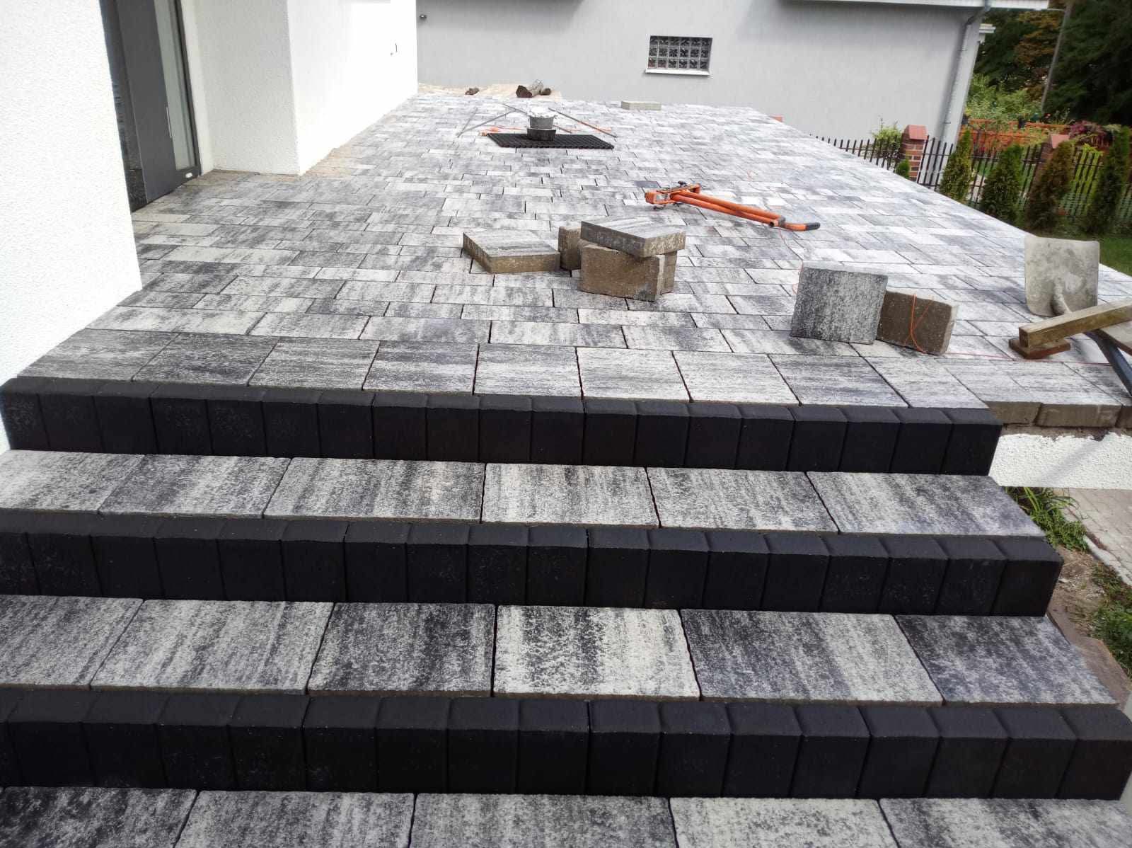 Schody i taras z kostki brukowej w odcieniach szarości, czarne obrzeża, częściowo ułożona nawierzchnia, narzędzia brukarskie widoczne na górze, dom w tle.