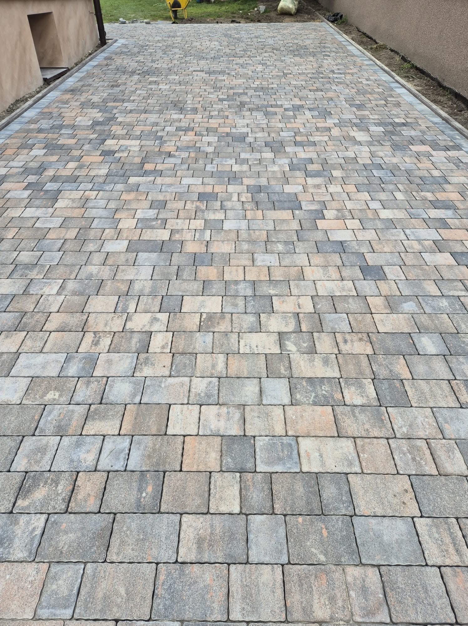 Szeroki kadr na ułożoną kostkę brukową w odcieniach szarości i beżu, tworzącą wzór na podjeździe. W tle widoczny budynek i fragment zieleni.