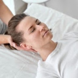 Kręgi Zdrowia Warszawa - Osteopatia - Fizjoterapia - Wkładki ortopedyczne - Kobieta leżąca na białym prześcieradle z zamkniętymi oczami, podczas gdy terapeuta delikatnie masuje jej szyję i głowę.
