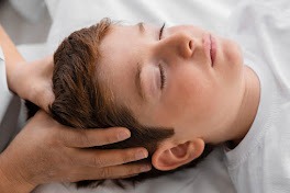 Chłopiec z zamkniętymi oczami leży na plecach, a dłonie terapeuty delikatnie podtrzymują jego głowę podczas sesji terapeutycznej.