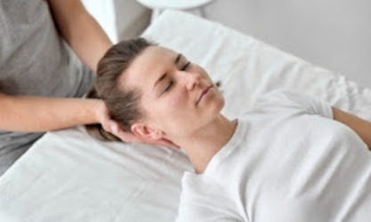 Kobieta leżąca na białym prześcieradle z zamkniętymi oczami, podczas gdy terapeuta delikatnie masuje jej szyję i głowę.