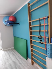 Sala do ćwiczeń z drabinką gimnastyczną, piłkami rehabilitacyjnymi na półce, matą do ćwiczeń na podłodze i gumami do ćwiczeń zawieszonymi na drabince.