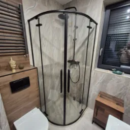 Nowoczesna łazienka z kabiną prysznicową z czarnymi profilami, marmurowe płytki na ścianach, wisząca toaleta i dekoracje w postaci złotej głowy Buddy oraz drewnianej szafki pod oknem z zieloną kaczką.