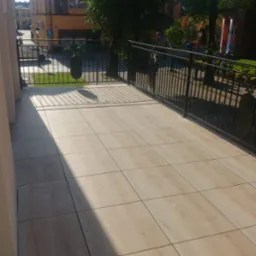 Jasne, drewnopodobne płytki na balkonie z czarną, metalową balustradą, widok na budynek i zieleń miejską w tle.