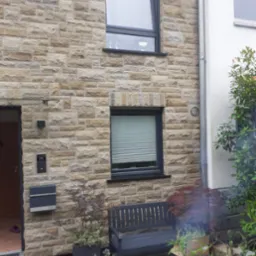 Elewacja budynku z piaskowca z szarą stolarką okienną, skrzynką na listy i ławką przed oknem z żaluzjami.