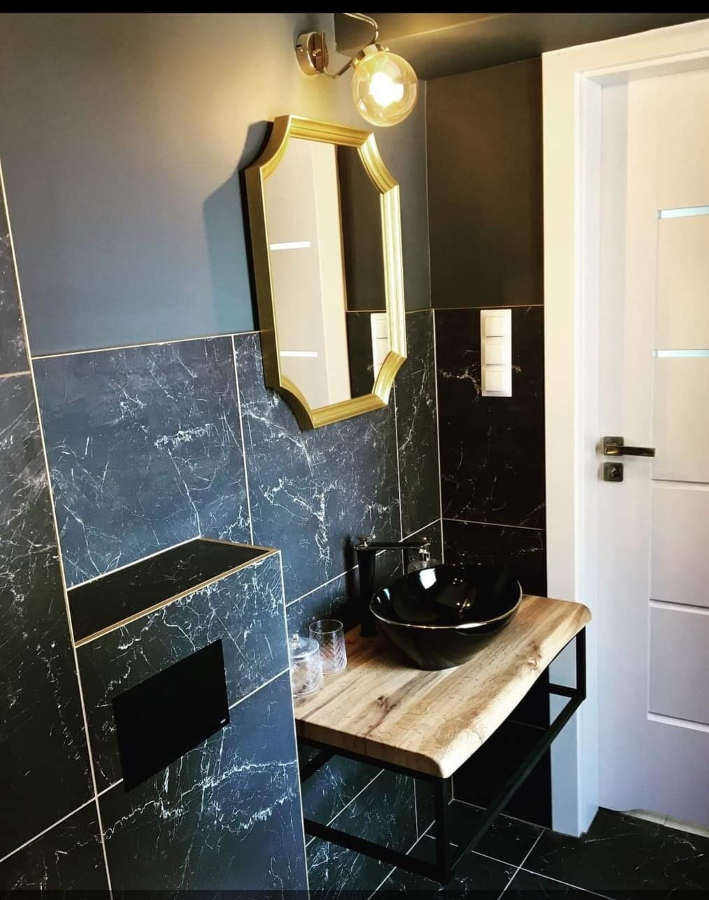 Nowoczesna łazienka z czarnymi płytkami imitującymi marmur, złotym lustrem i umywalką nablatową na drewnianym blacie z metalową podstawą.