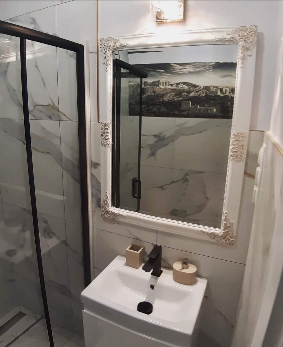 Umywalka z czarną baterią i ozdobnym lustrem w łazience wykończonej płytkami imitującymi marmur, z widoczną kabiną prysznicową.