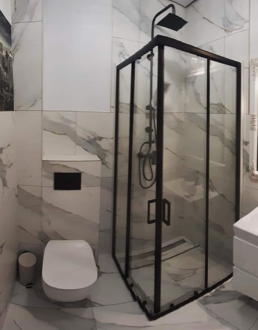 Nowoczesna łazienka z kabiną prysznicową w czarnej ramie, marmurowe płytki na ścianach i podłodze, wisząca toaleta z czarnym przyciskiem spłukującym.