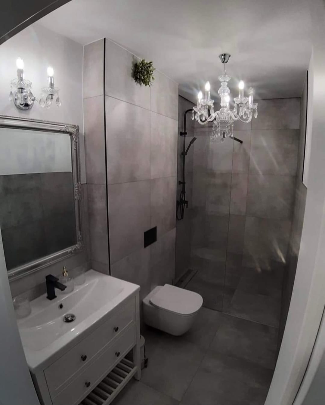 Wykończona łazienka w szarościach z kryształowym żyrandolem, umywalką na szafce z czarną armaturą i wiszącą toaletą.