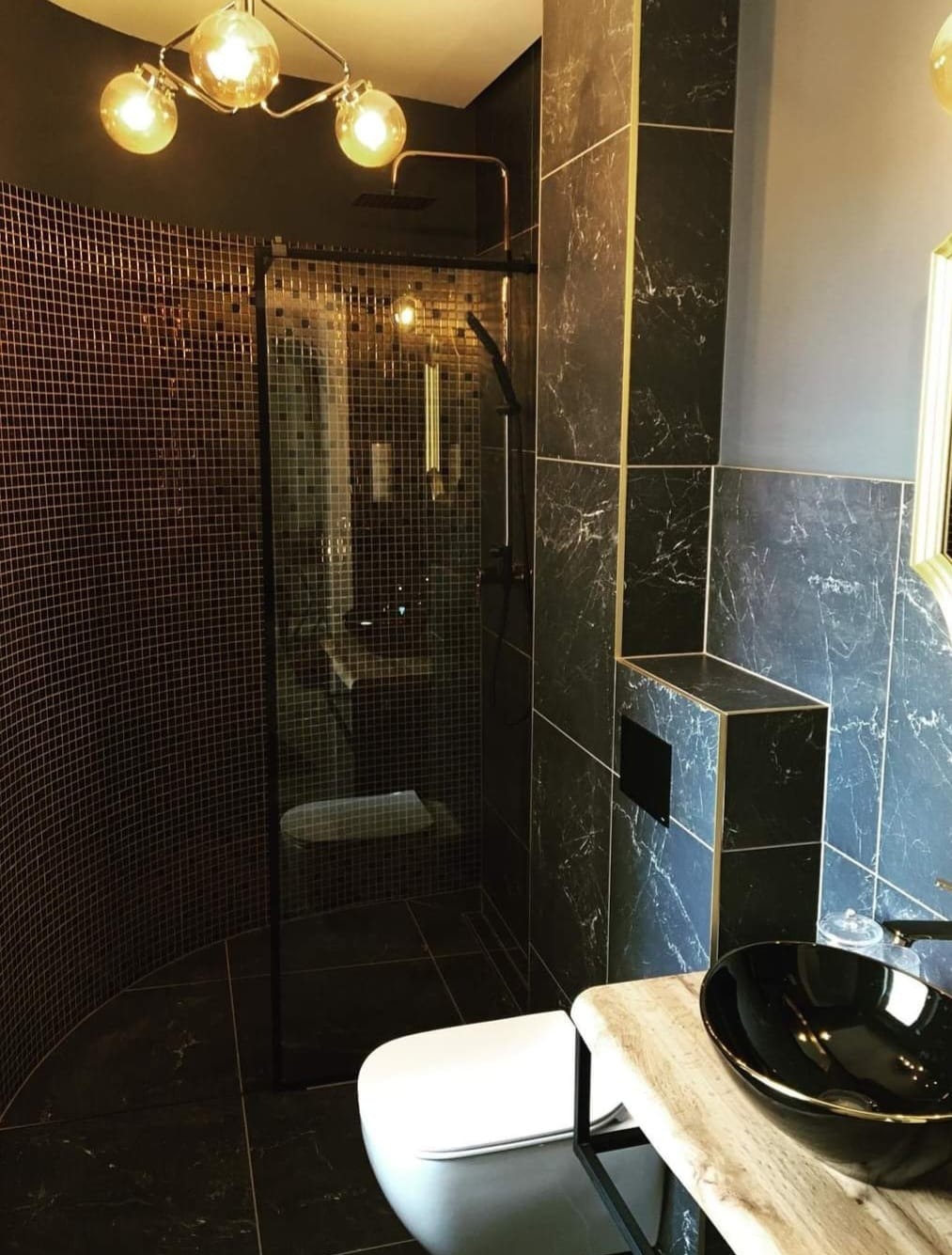 Nowoczesna łazienka w Oświęcimiu z kabiną prysznicową wyłożoną mozaiką, czarnymi płytkami imitującymi marmur z złotymi akcentami, umywalką w czarnej misie i designerską lampą sufitową.