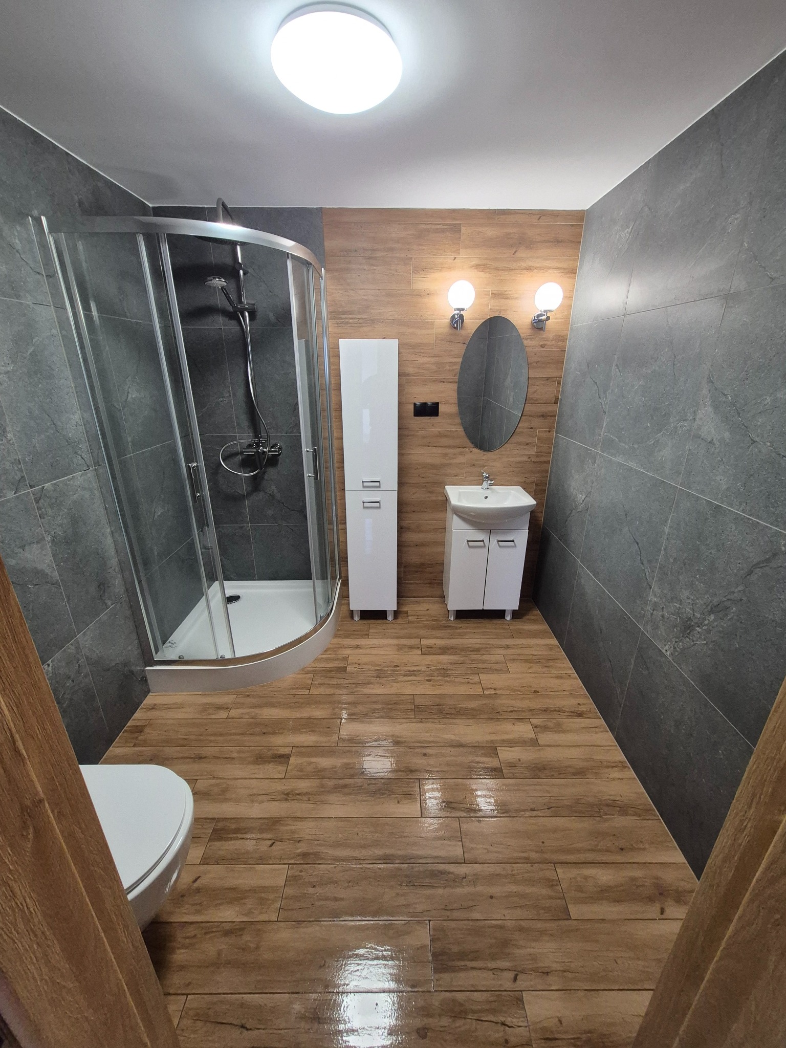 Nowoczesna łazienka z kabiną prysznicową, umywalką z szafką i wysoką białą szafką, wykończona szarymi płytkami imitującymi kamień i drewnopodobnymi płytkami na podłodze.