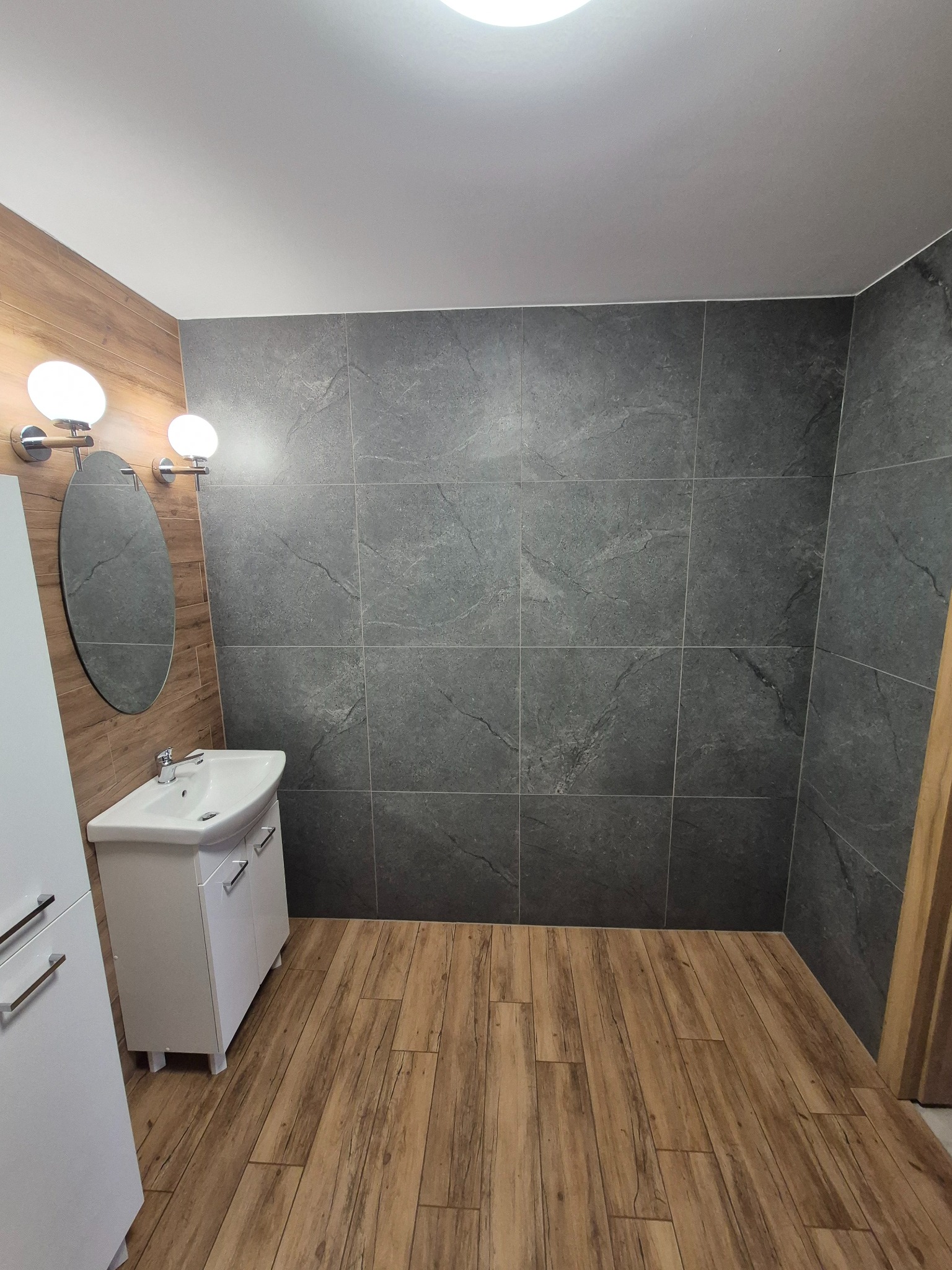 Wykończona łazienka z szarymi płytkami imitującymi kamień na ścianach, drewnopodobną podłogą, białą szafką umywalkową i owalnym lustrem z dwoma okrągłymi lampami.