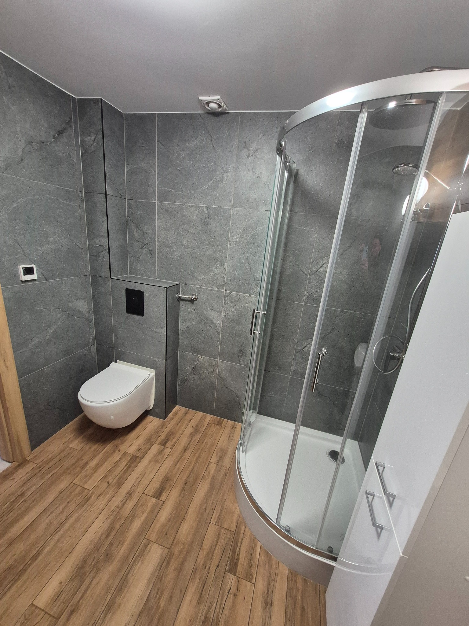 Nowoczesna łazienka z szarymi płytkami imitującymi kamień, drewnopodobną podłogą, kabiną prysznicową z zaokrąglonym frontem i wiszącą toaletą z czarnym przyciskiem.