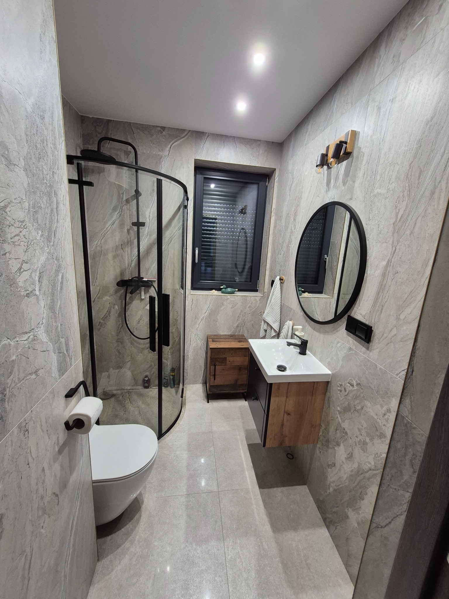 Nowoczesna łazienka z szarymi marmurkowymi płytkami, kabiną prysznicową z czarnymi profilami, umywalką z szafką w kolorze drewna i okrągłym lustrem z czarną ramą.