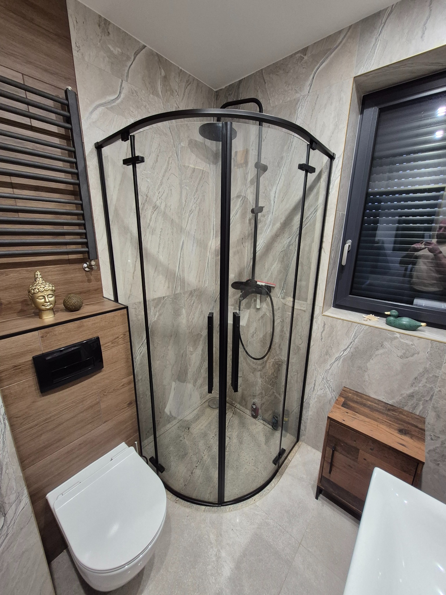 Nowoczesna łazienka z kabiną prysznicową z czarnymi profilami, marmurowe płytki na ścianach, wisząca toaleta i dekoracje w postaci złotej głowy Buddy oraz drewnianej szafki pod oknem z zieloną kaczką.