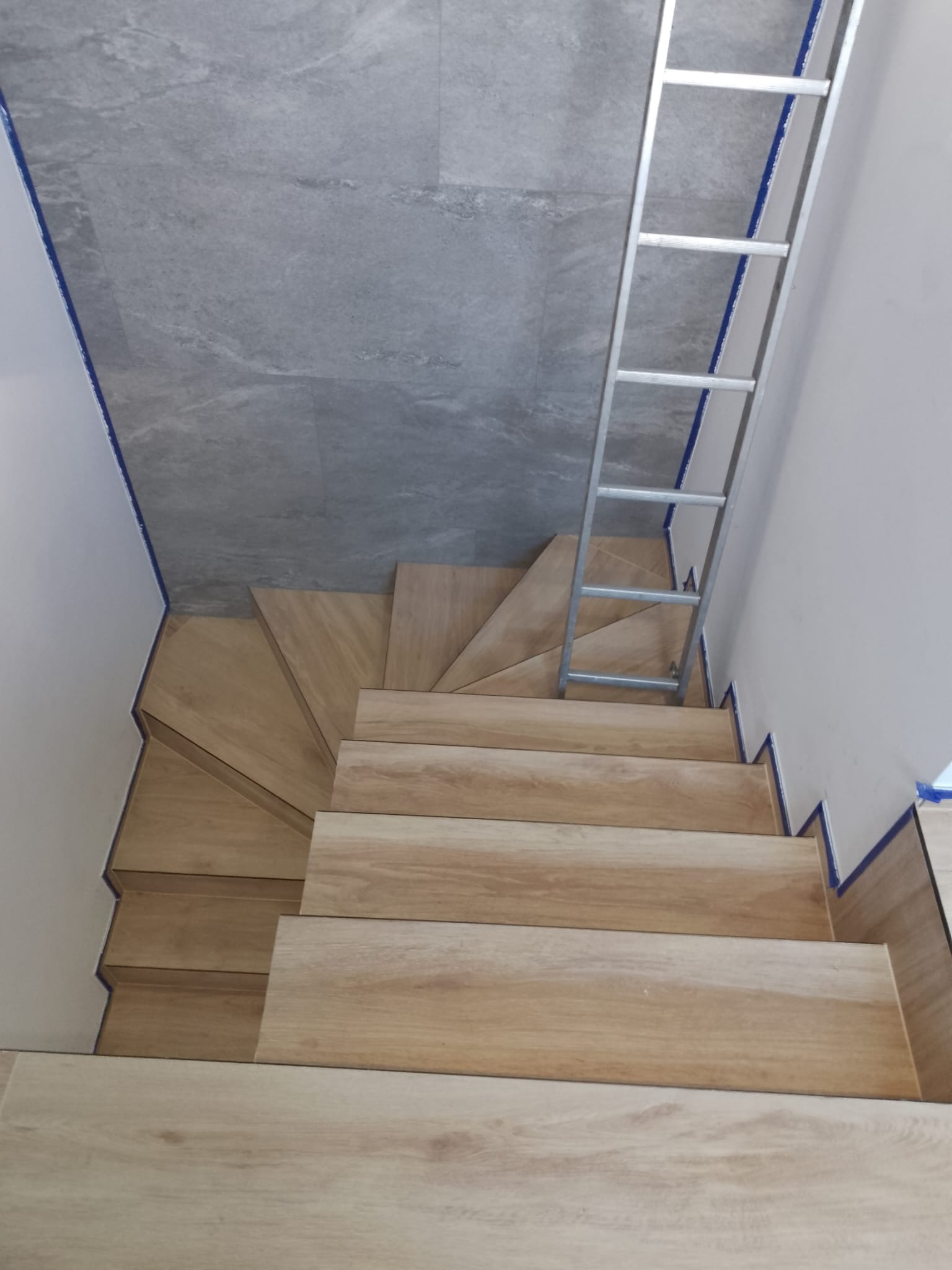 Klatka schodowa wyłożona płytkami imitującymi drewno, widok z góry, z metalową drabiną opartą o ścianę i niebieską taśmą malarską zabezpieczającą krawędzie.