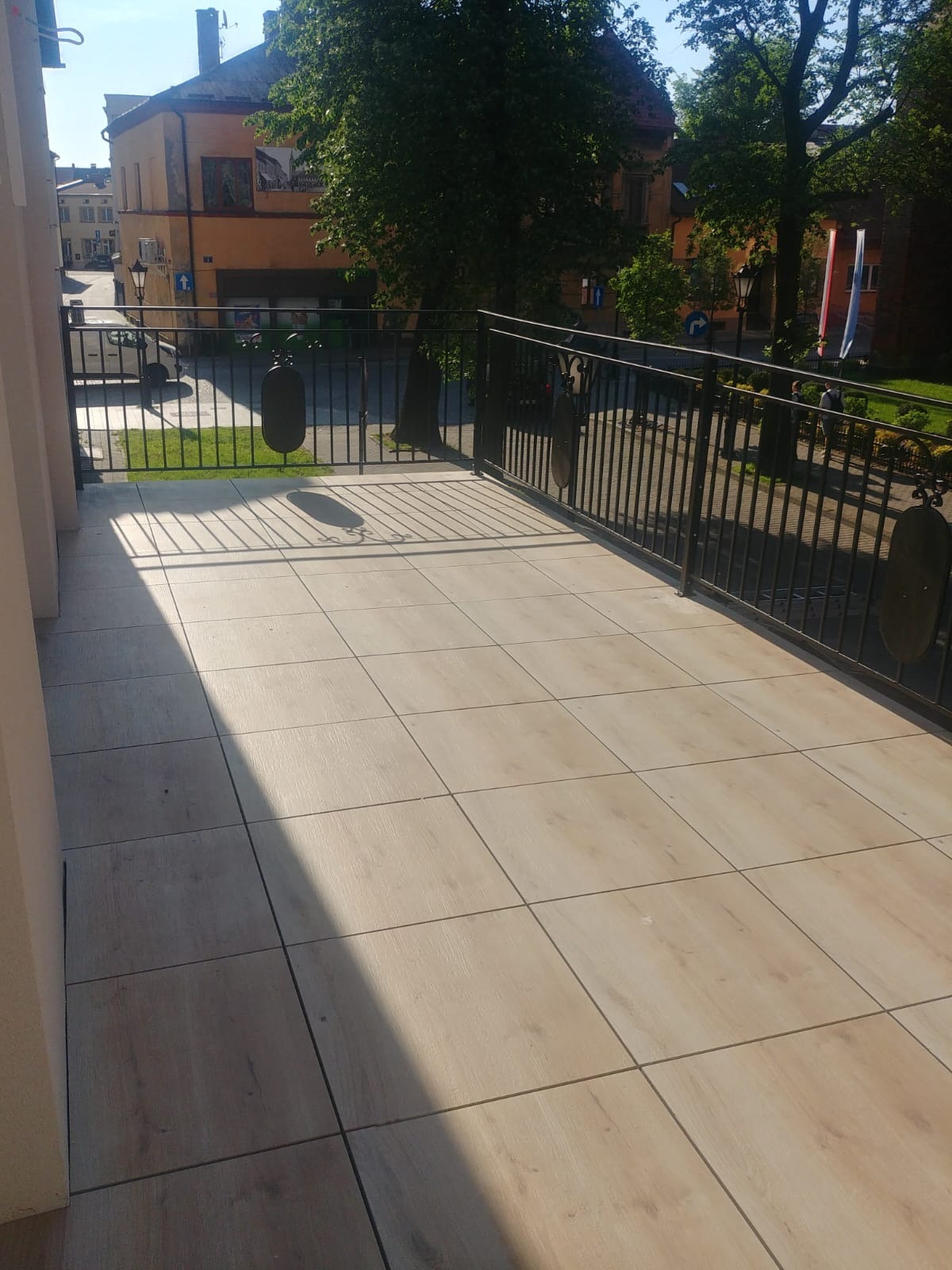 Jasne, drewnopodobne płytki na balkonie z czarną, metalową balustradą, widok na budynek i zieleń miejską w tle.