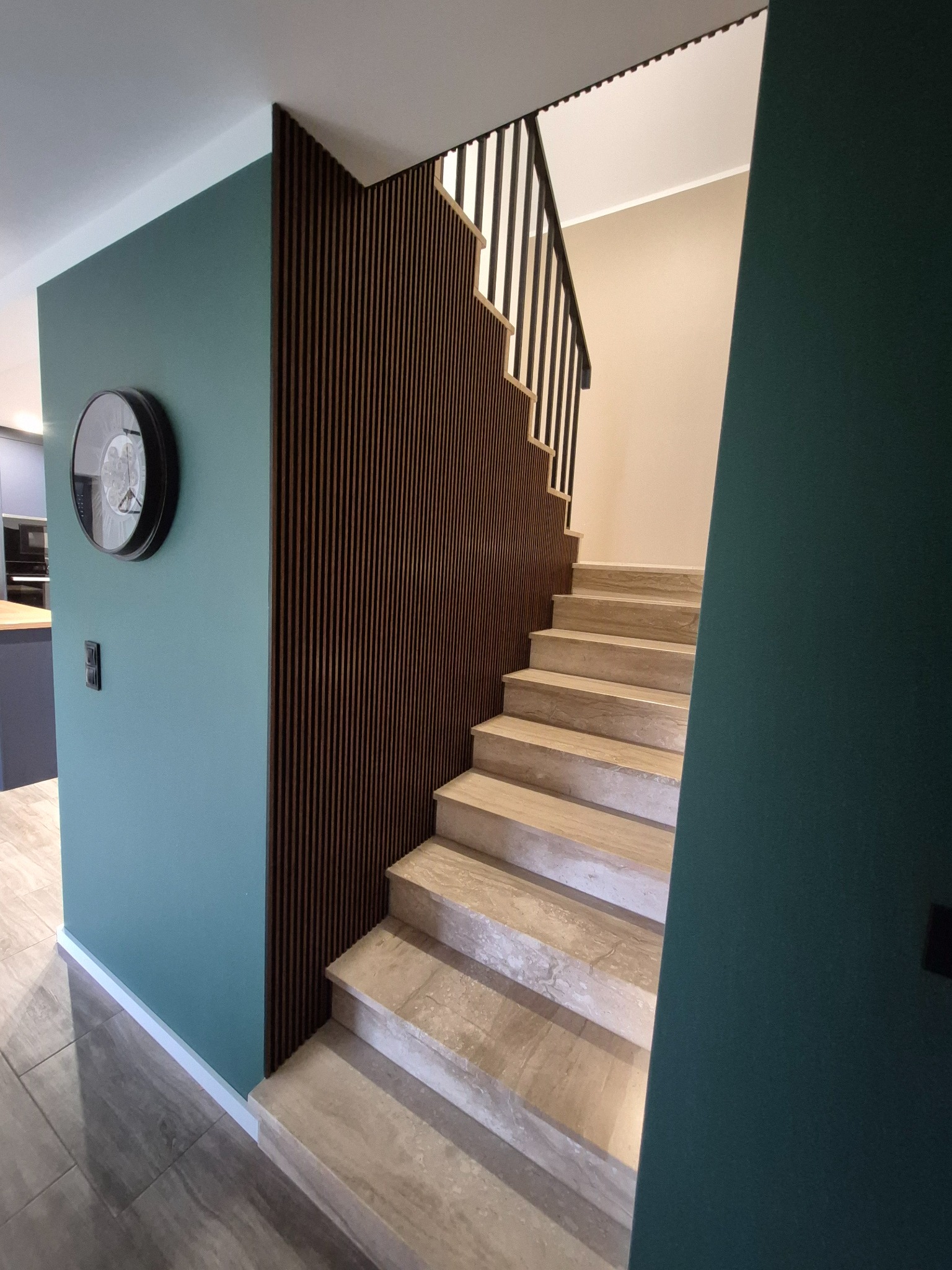 Nowoczesne schody z trawertynu z czarną balustradą, obok ściana w kolorze butelkowej zieleni z okrągłym zegarem i dekoracyjną ścianką z drewnianych lameli.