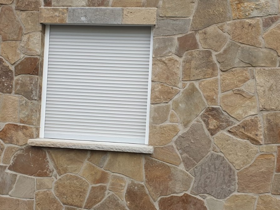 Zamknięta, biała roleta okienna na tle ściany z nieregularnych, brązowo-beżowych kamieni.