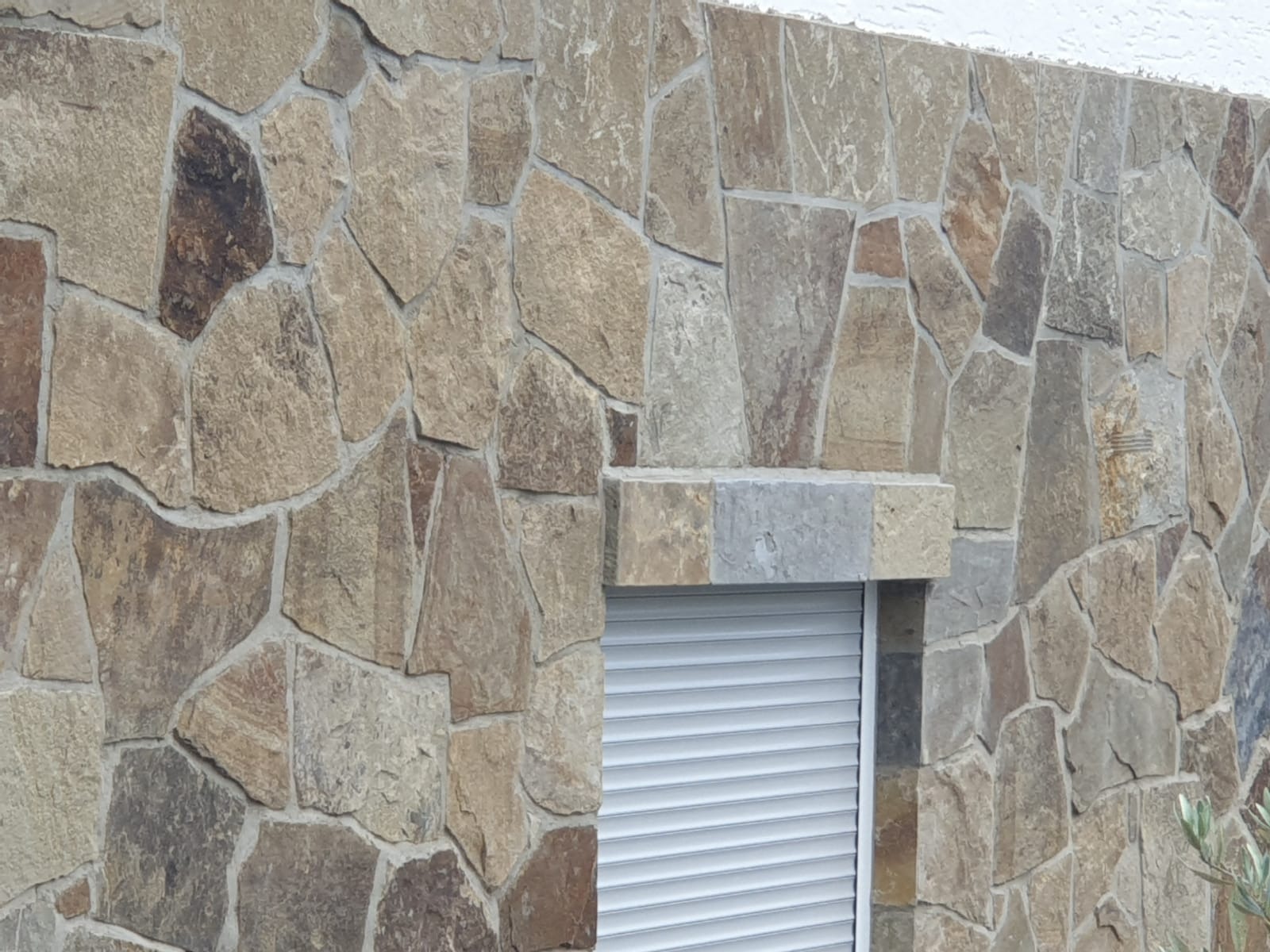 Elewacja budynku wykończona nieregularnym kamieniem w odcieniach beżu i brązu, z widocznymi szarymi spoinami, okno z opuszczoną białą roletą.