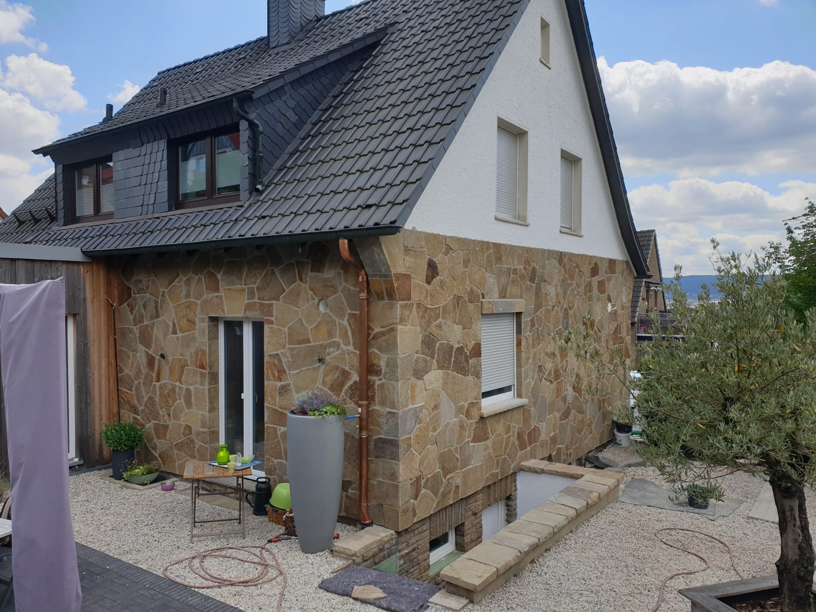 Elewacja domu wykończona nieregularnym kamieniem w odcieniach beżu i brązu, z widocznymi oknami z białymi roletami i fragmentem dachu pokrytego ciemną dachówką.