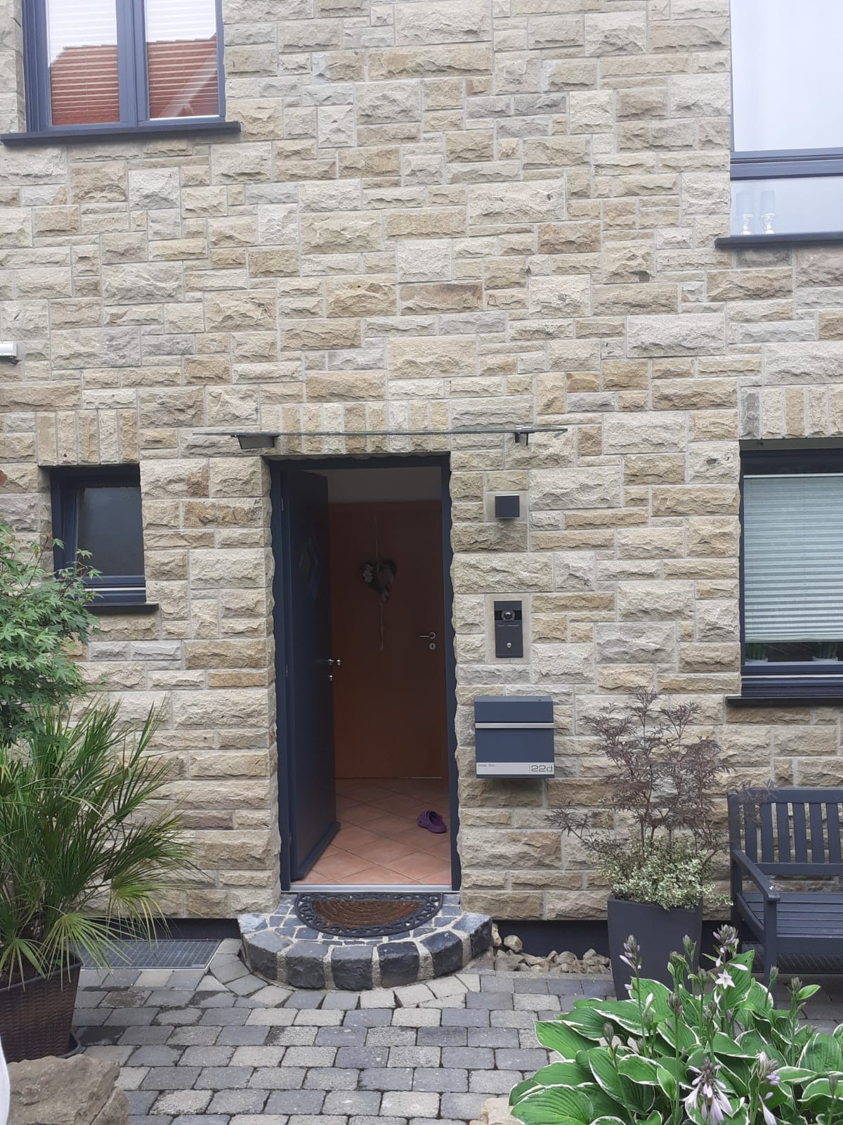 Elewacja domu z piaskowca z otwartymi, grafitowymi drzwiami wejściowymi i skrzynką pocztową 22d, widok na wnętrze z wycieraczką i parą fioletowych kapci.