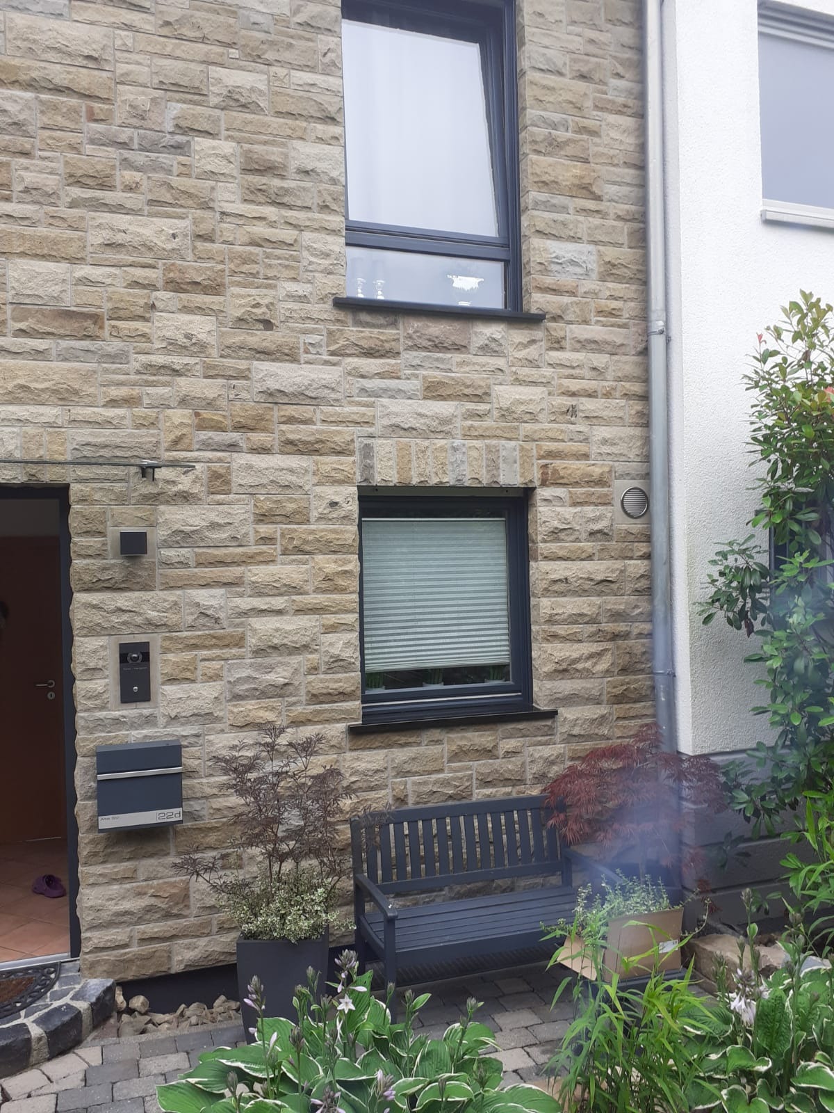 Elewacja budynku z piaskowca z szarą stolarką okienną, skrzynką na listy i ławką przed oknem z żaluzjami.