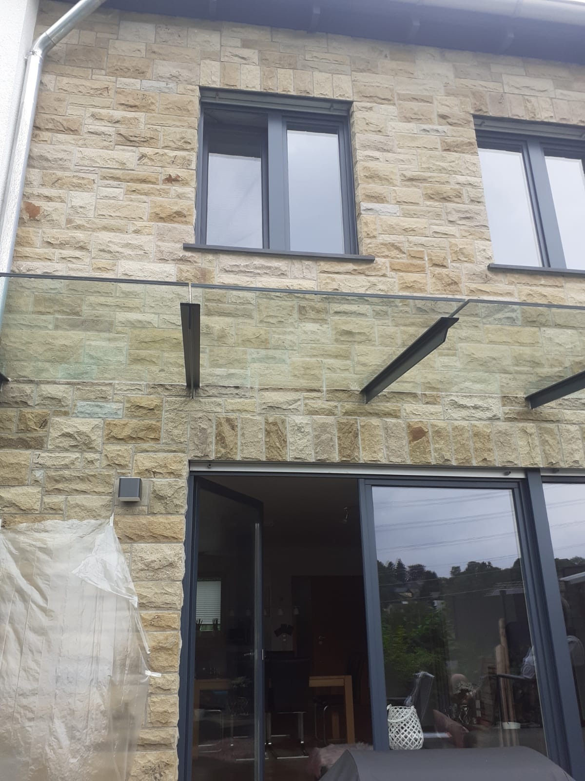 Elewacja domu z kamienia naturalnego z szarą stolarką okienną i szklanym zadaszeniem nad tarasem, zabezpieczona folią malarską.