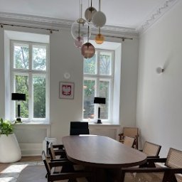 KANCELARIA NOTARIALNA ZINE ABASS ALI - Sala konferencyjna z owalnym drewnianym stołem i krzesłami z rattanowym oparciem w kancelarii w Warszawie. Widoczne duże okna z widokiem na drzewa, żyrandol z szklanych kul i godło Polski na ścianie.