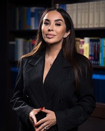 Portret kobiety w czarnym, dwurzędowym garniturze na tle regału z książkami. Dłonie splecione przed sobą, delikatny uśmiech.