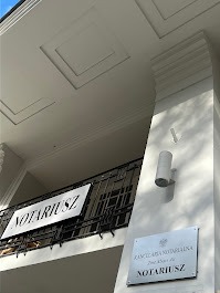 Elewacja budynku z szyldami informującymi o Kancelarii Notarialnej; widok z dołu, z lekkim pochyleniem w lewo, ukazujący balkon z czarną, ozdobną balustradą i tabliczkę z nazwą kancelarii.